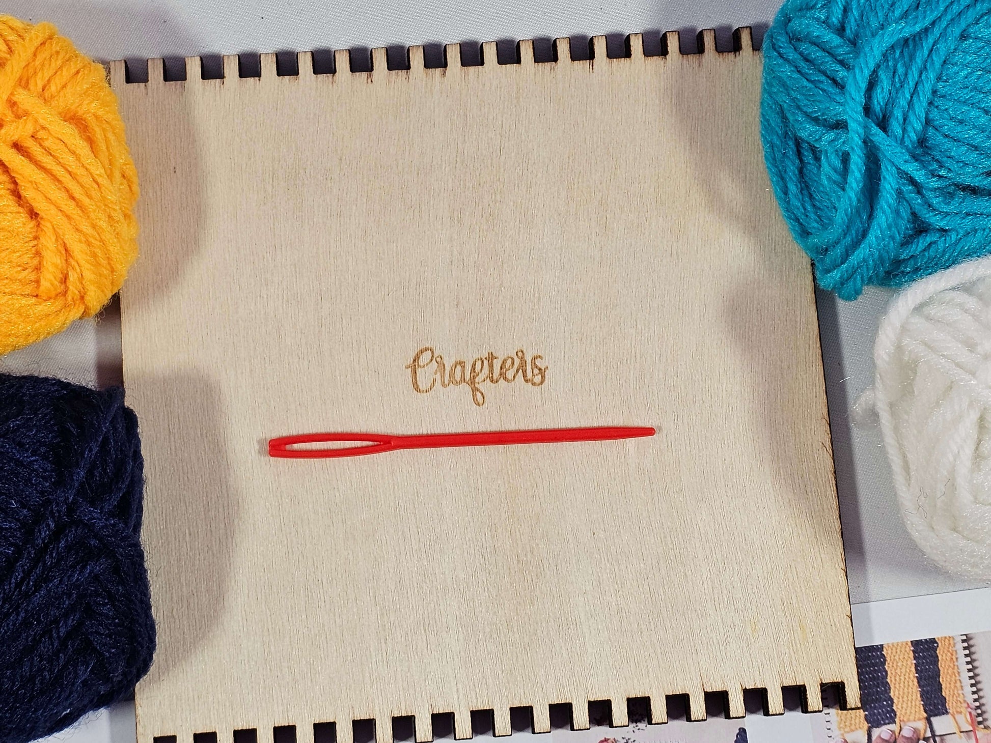 Crafters Loom Coaster Kit with wooden loom, red needle, and yarn balls in dark blue, light blue, yellow, and white.