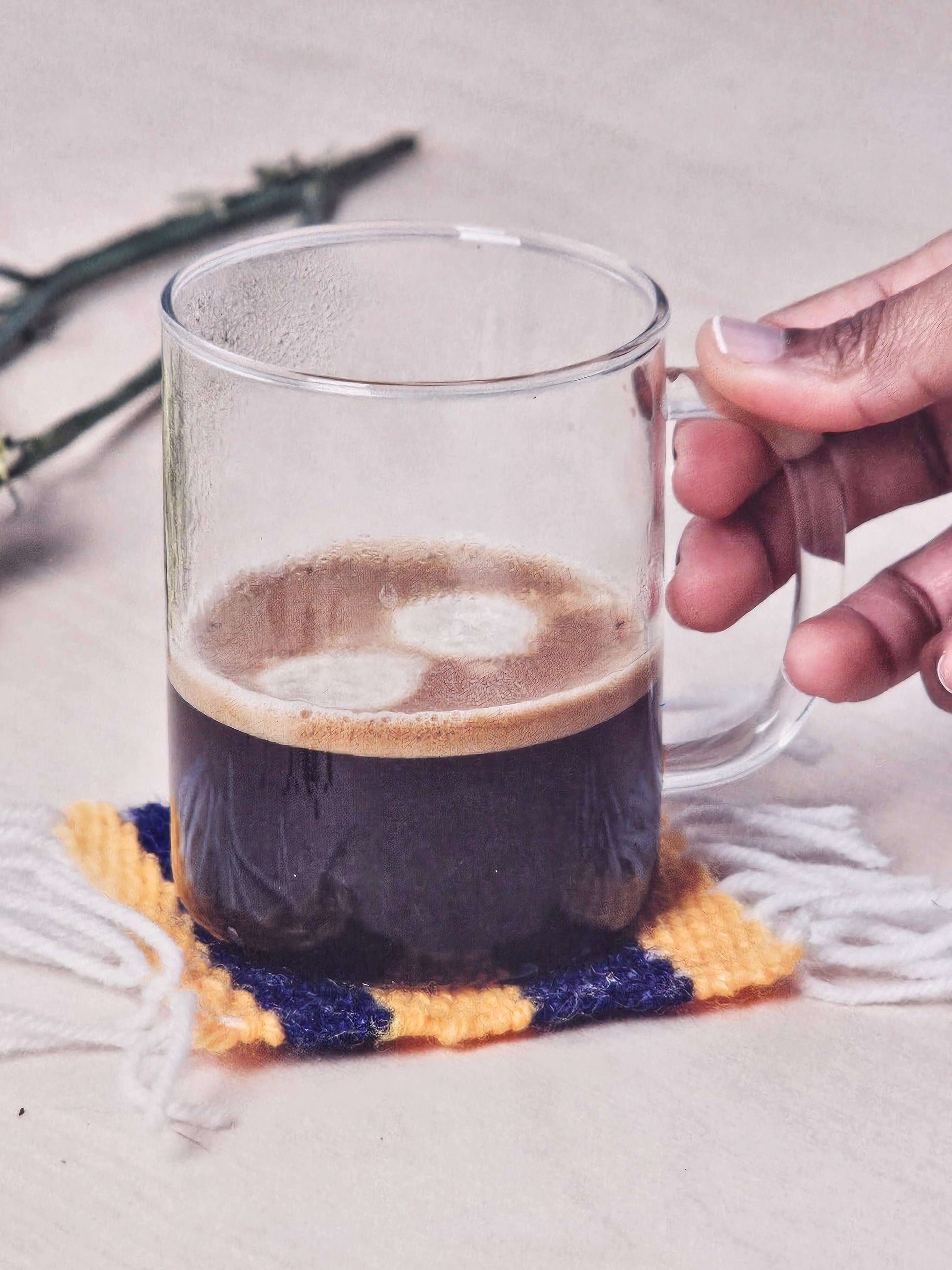 Handcrafted yellow and blue coaster holding a glass of coffee, showcasing the Crafters Loom Coaster Kit.