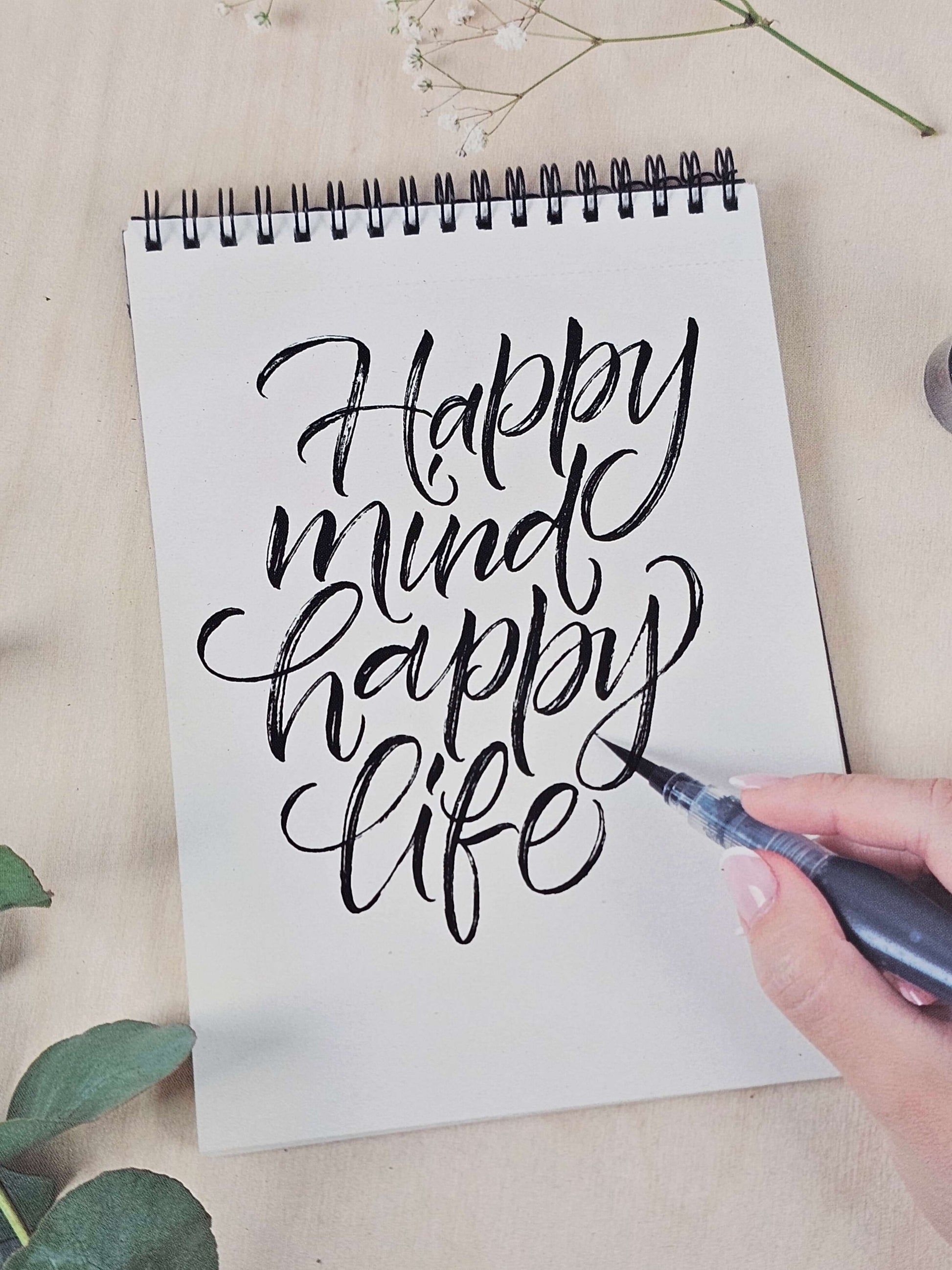 Hand lettering practice showing the phrase 'Happy mind, happy life' in a sketchbook surrounded by greenery.