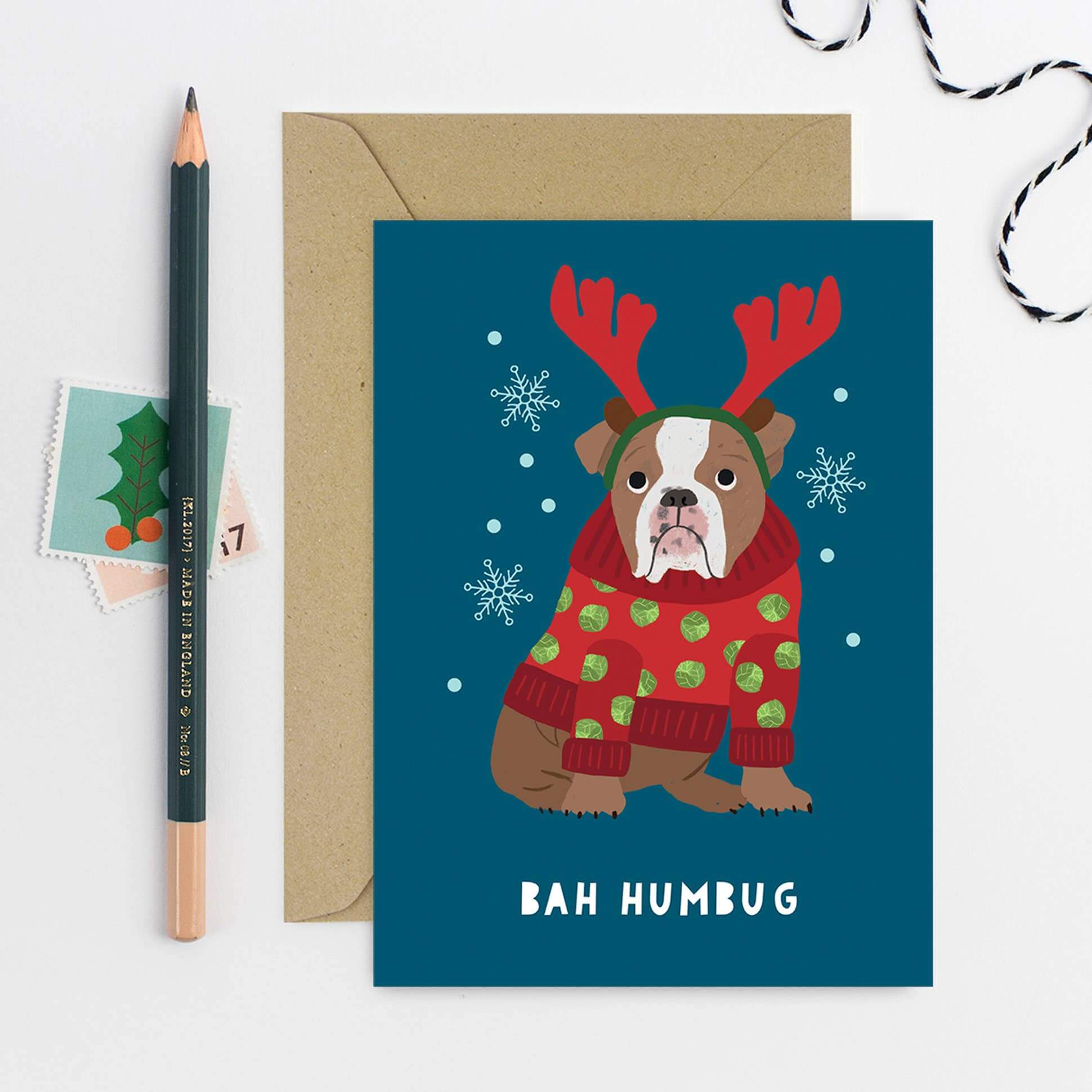 Christmas card with a dog wearing reindeer antlers and a sweater, next to an envelope and pencil.