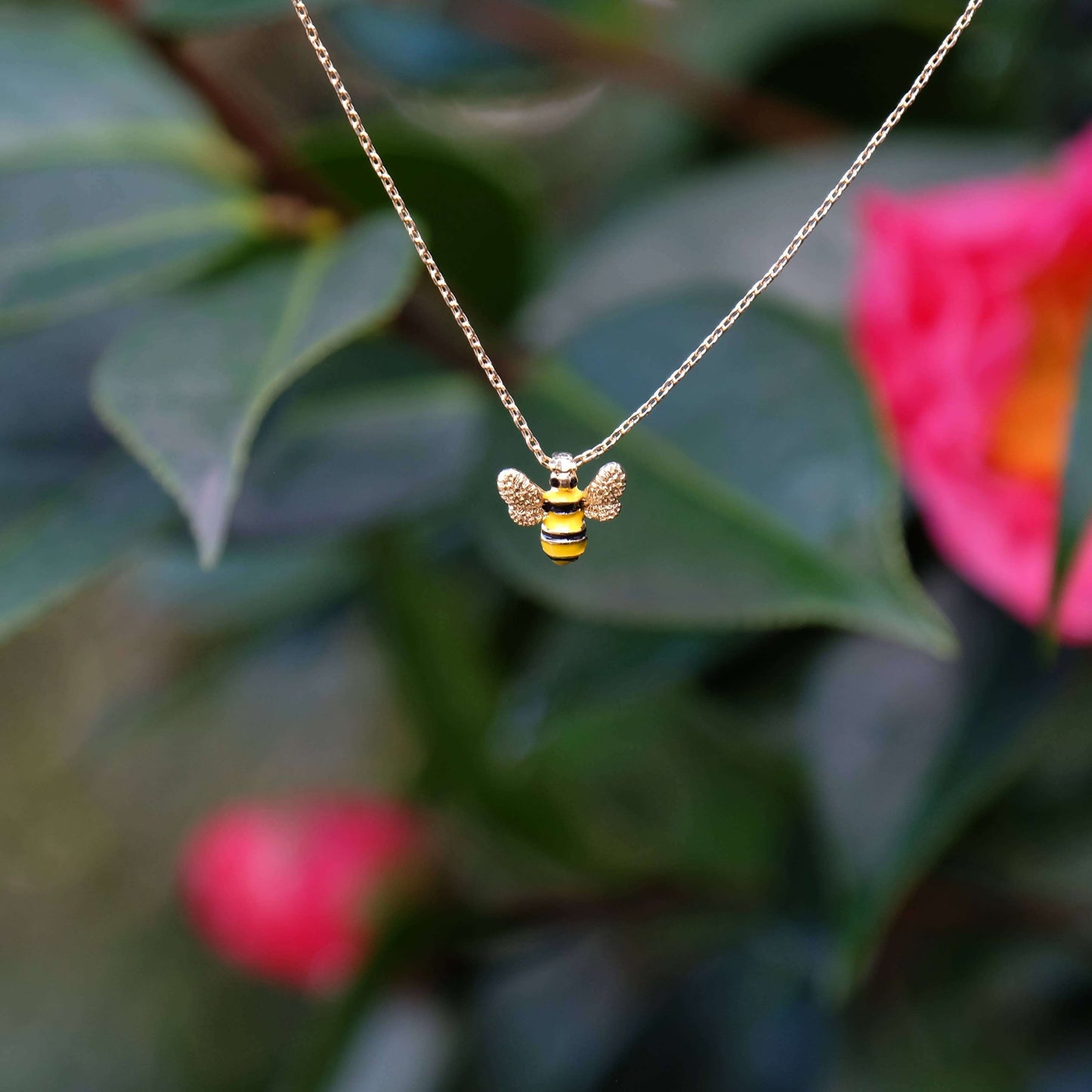 Gold bee-shaped pendant necklace on a chain against a blurred natural background with pink flowers and green leaves.
