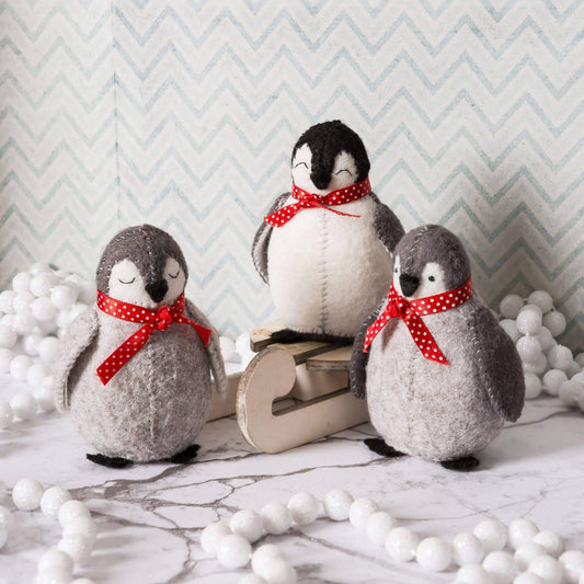 Three felt penguins with red polka dot bows on a marble surface with decorative balls and a chevron-patterned wall.