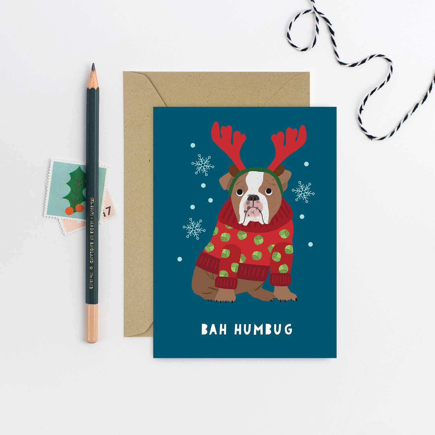 Christmas card with a dog wearing a sweater and reindeer antlers on a blue background, with an envelope and stationery items.