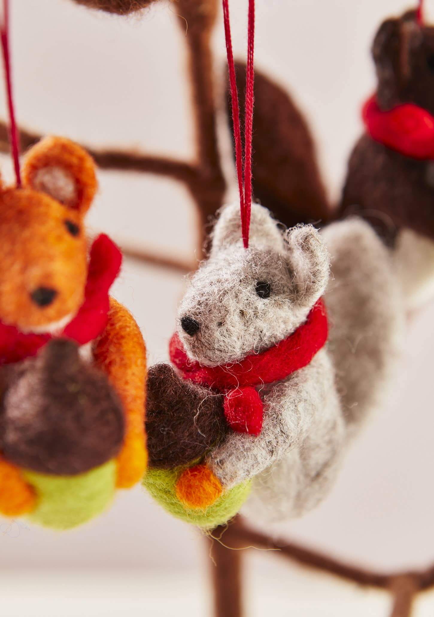 Felted animal ornaments, including a fox and a deer, hanging with red scarves against a blurred background.