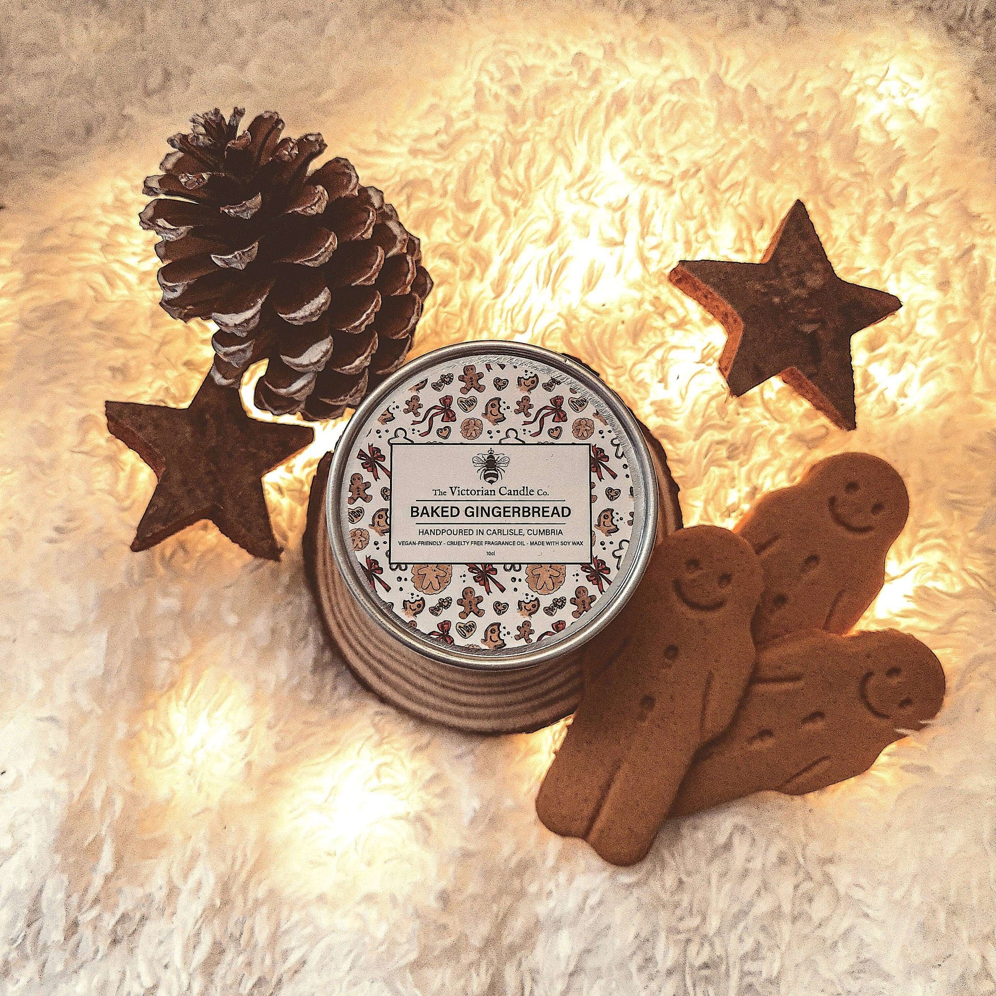 Candle labeled 'Baked Gingerbread' surrounded by decorative gingerbread men, stars, and a pine cone on a soft surface.