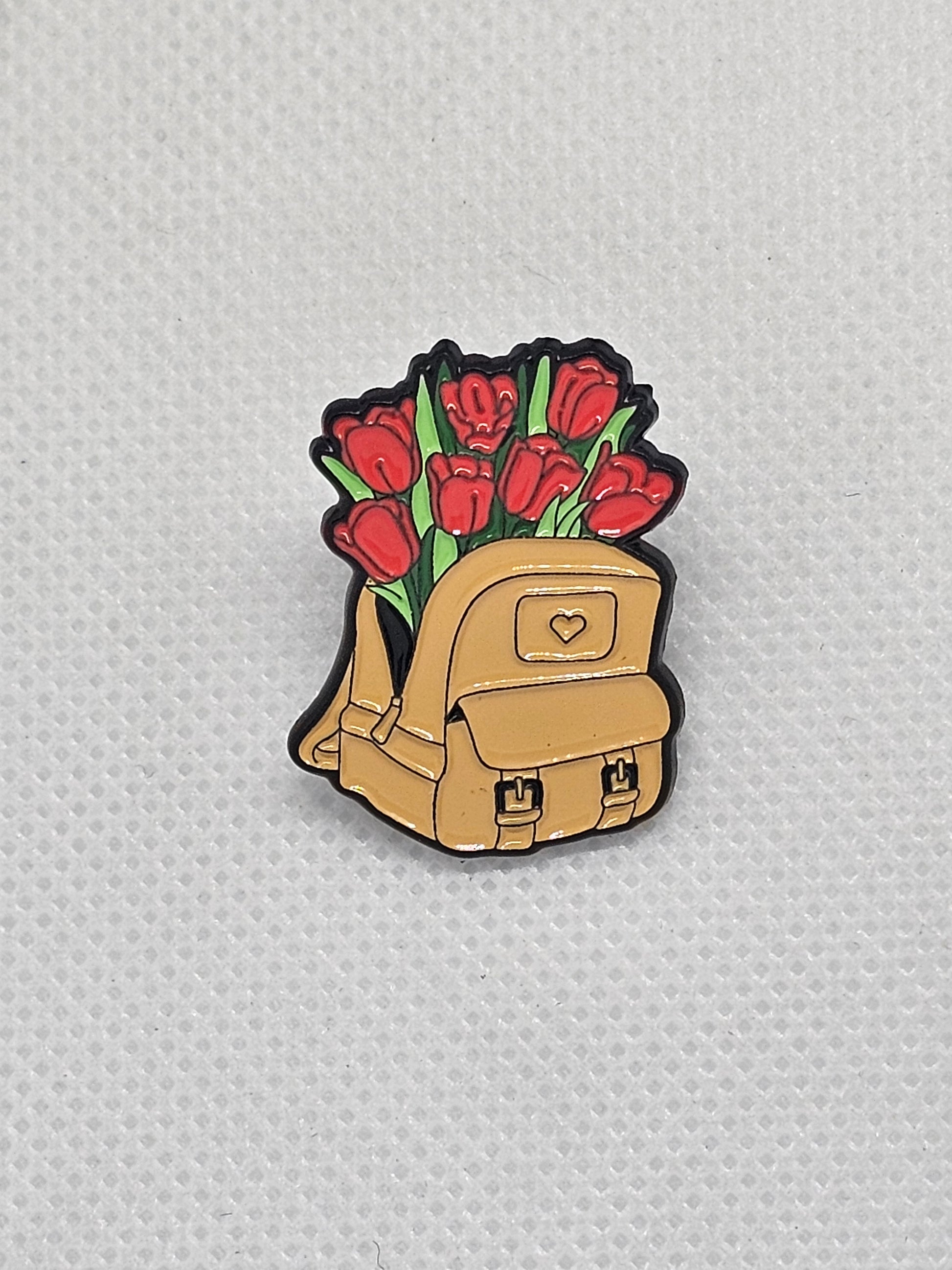 Enamel pin badge featuring a cute backpack with red tulips for plant lovers. Perfect accessory for any nature enthusiast.