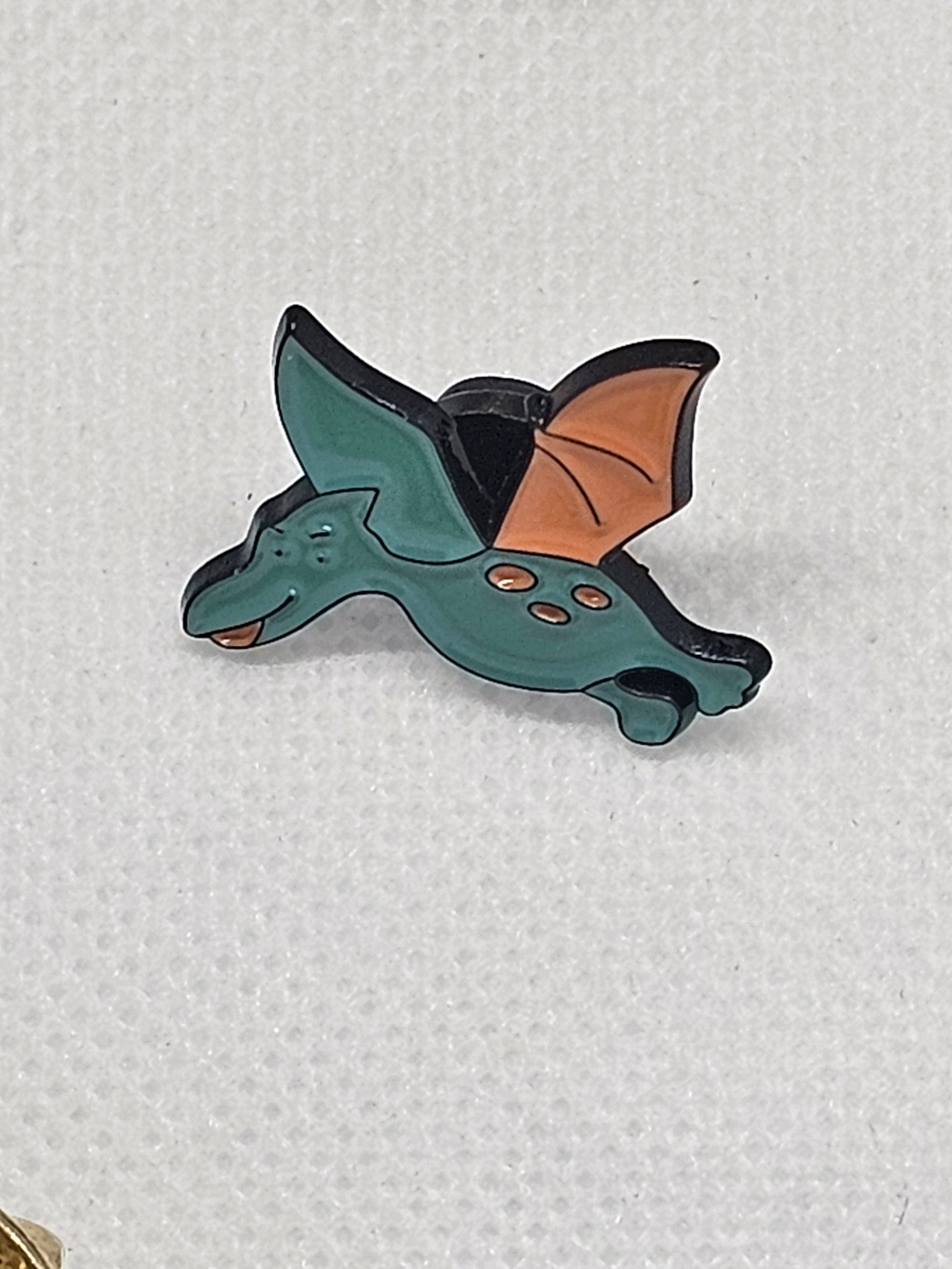 Cute flying dinosaur enamel pin badge in teal and orange, perfect for bags or clothing accessories.