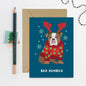 Christmas card with a dog wearing reindeer antlers and a sweater, next to an envelope and pencil.