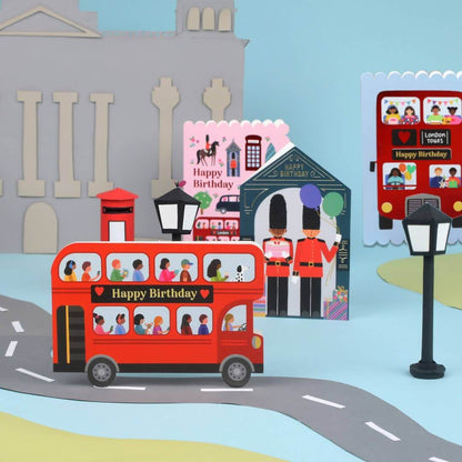 Illustration of a red double-decker bus with people on a road, surrounded by London-themed items like a postbox and a birthday card.