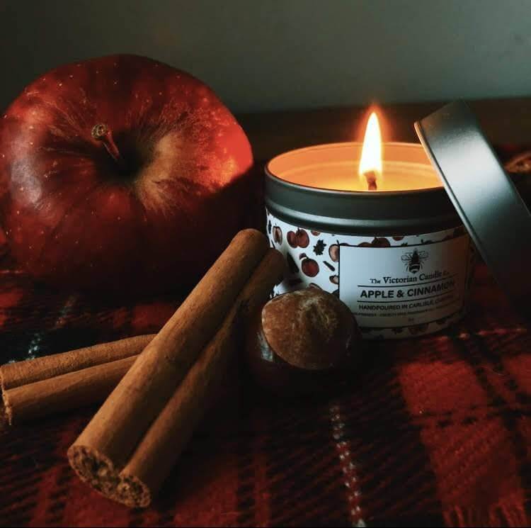 Candle labeled 'Apple & Cinnamon' with an apple, cinnamon sticks, and a nut on a dark surface.