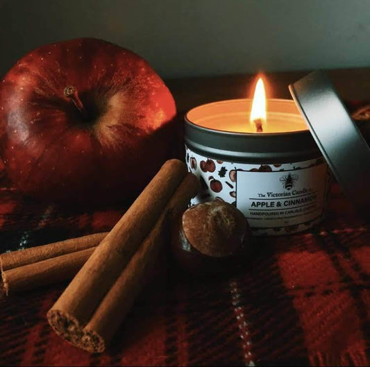 Candle labeled 'Apple & Cinnamon' with an apple, cinnamon sticks, and a nut on a dark surface.
