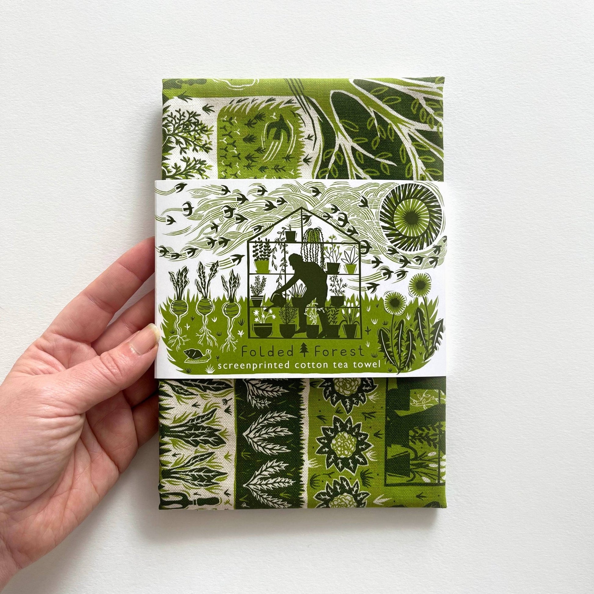 Hand holding a green and white patterned tea towel with 'Folded Forest' branding.
