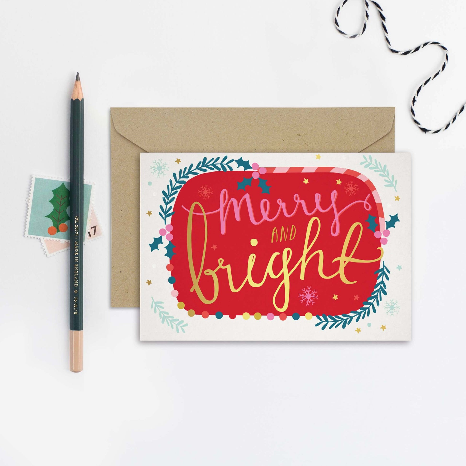 Christmas card with 'Merry and Bright' text, envelope, pencil, and decorative string on a white background
