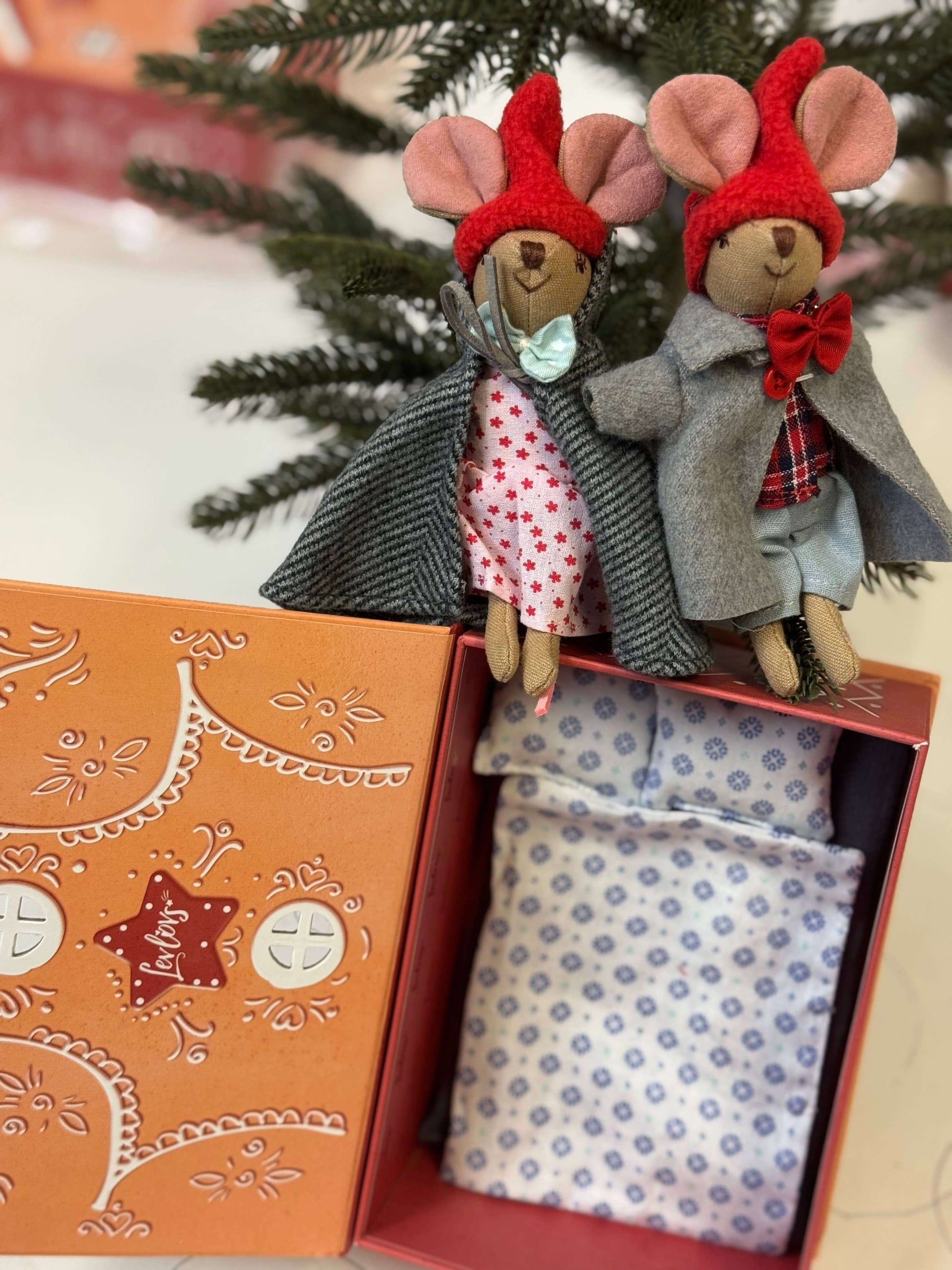 Two small mouse figurines in winter clothing on a decorative box with a Christmas tree in the background.