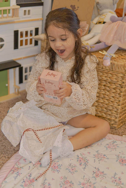 Young girl in a white dress sitting on a floral bedspread holding a pink box.