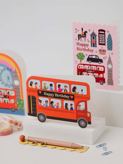 Card with a red double-decker bus design and 'Happy Birthday' text, surrounded by other cards and stationery items.