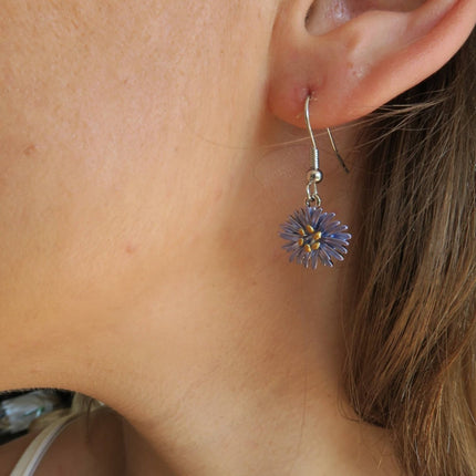 A close-up of a person's ear wearing a dangling flower earring. The earring has a silver-toned fishhook back and a small purple flower charm with a yellow center, shown against skin and brown hair.