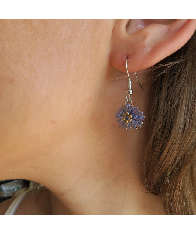 A close-up of a person's ear wearing a dangling flower earring. The earring has a silver-toned fishhook back and a small purple flower charm with a yellow center, shown against skin and brown hair.