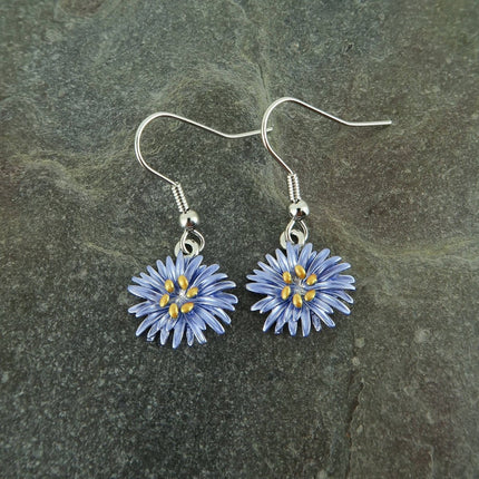 A pair of handmade dangle earrings featuring small blue and yellow flower charms on silver fishhook wires, displayed on a dark textured slate surface.
