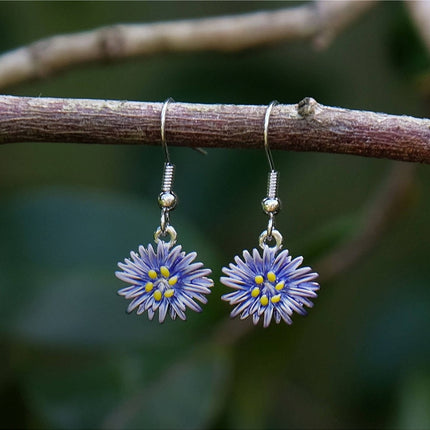 A pair of dangling earrings shaped like small purple flowers with yellow centers, hanging from silver hooks on a brown tree branch against a blurred green background.