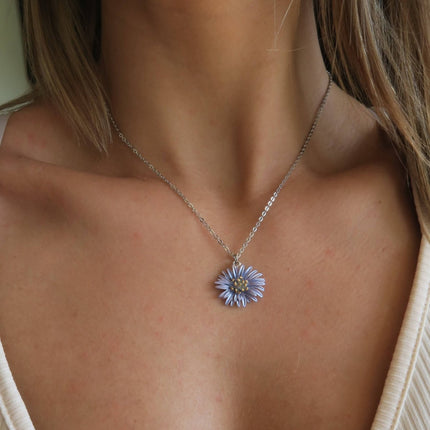 A close-up of a person's neck and chest wearing a delicate silver chain necklace with a small lavender-purple flower pendant.