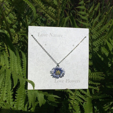 A silver necklace with a blue and yellow flower pendant displayed on a white card that reads 'Love Nature' and 'Love Flowers', set against a background of green ferns.