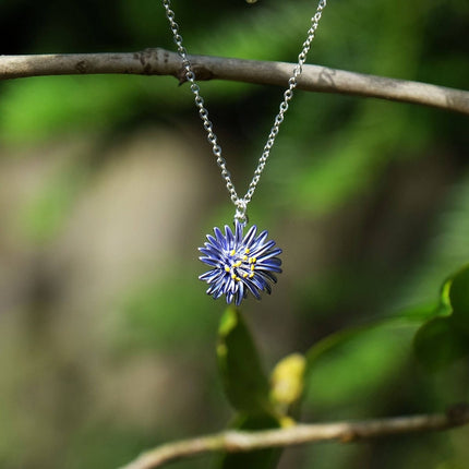 A silver chain necklace with a flower-shaped pendant featuring blue petals and a yellow center, hanging from a tree branch against a blurred green natural background.