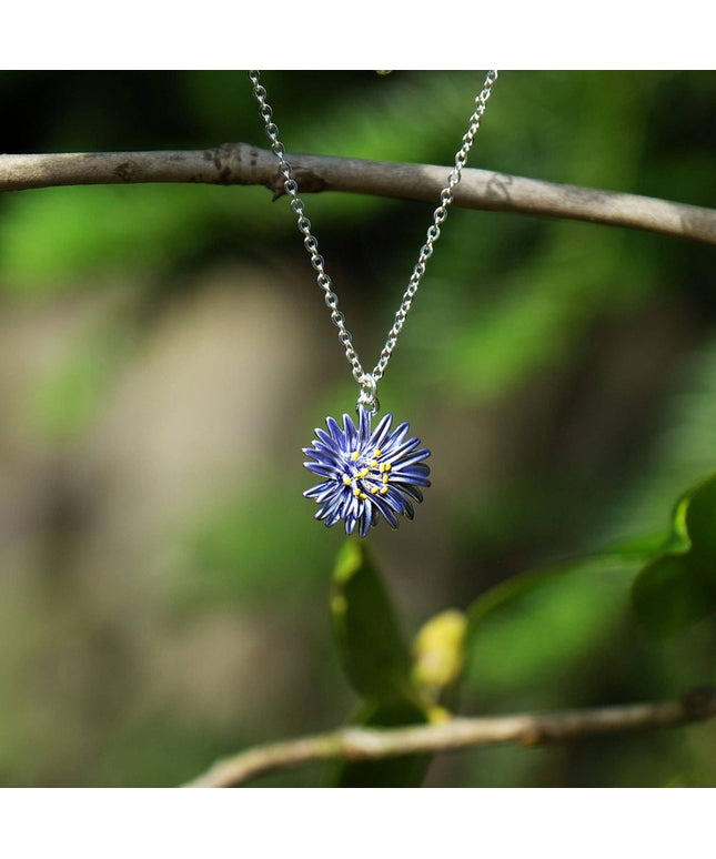 A silver chain necklace with a flower-shaped pendant featuring blue petals and a yellow center, hanging from a tree branch against a blurred green natural background.