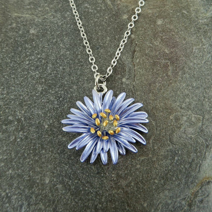 A silver chain necklace with a daisy flower pendant, featuring light blue petals and a gold center, displayed on a textured gray stone background.