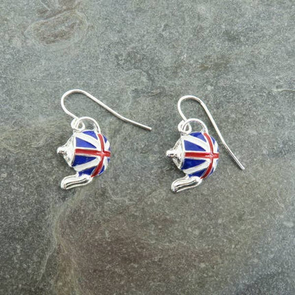 A pair of silver fishhook earrings with teapot-shaped charms decorated in a red, white, and blue Union Jack pattern, displayed on a textured gray slate surface.