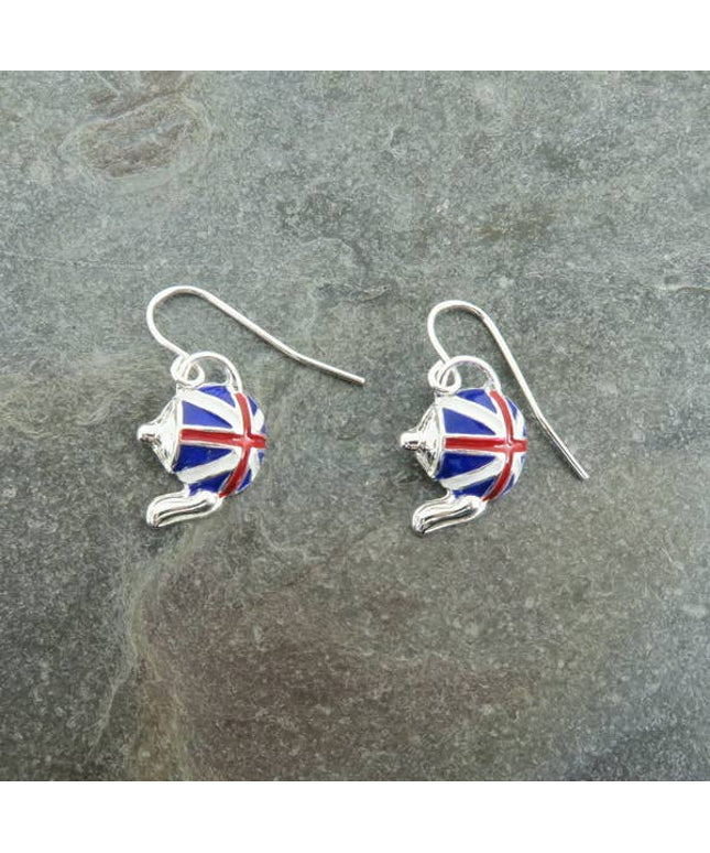 A pair of silver fishhook earrings with teapot-shaped charms decorated in a red, white, and blue Union Jack pattern, displayed on a textured gray slate surface.