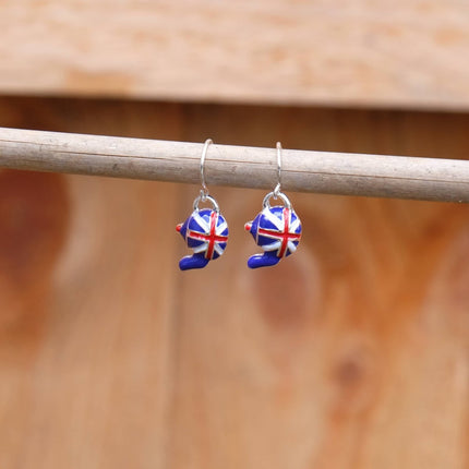 A pair of silver-toned dangle earrings with small enamel bear charms decorated in a red, white, and blue Union Jack pattern, hanging from a horizontal wooden rod against a warm, blurred wood background.