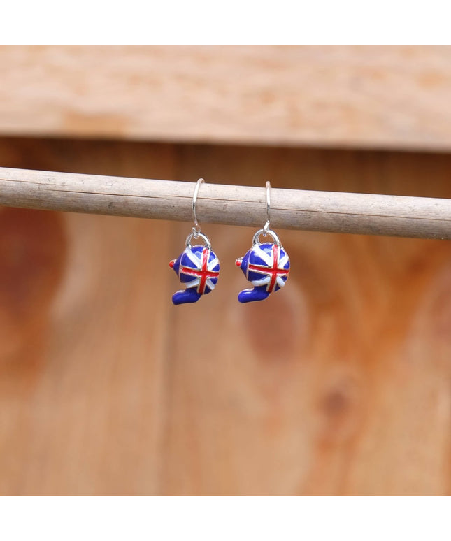 A pair of silver-toned dangle earrings with small enamel bear charms decorated in a red, white, and blue Union Jack pattern, hanging from a horizontal wooden rod against a warm, blurred wood background.