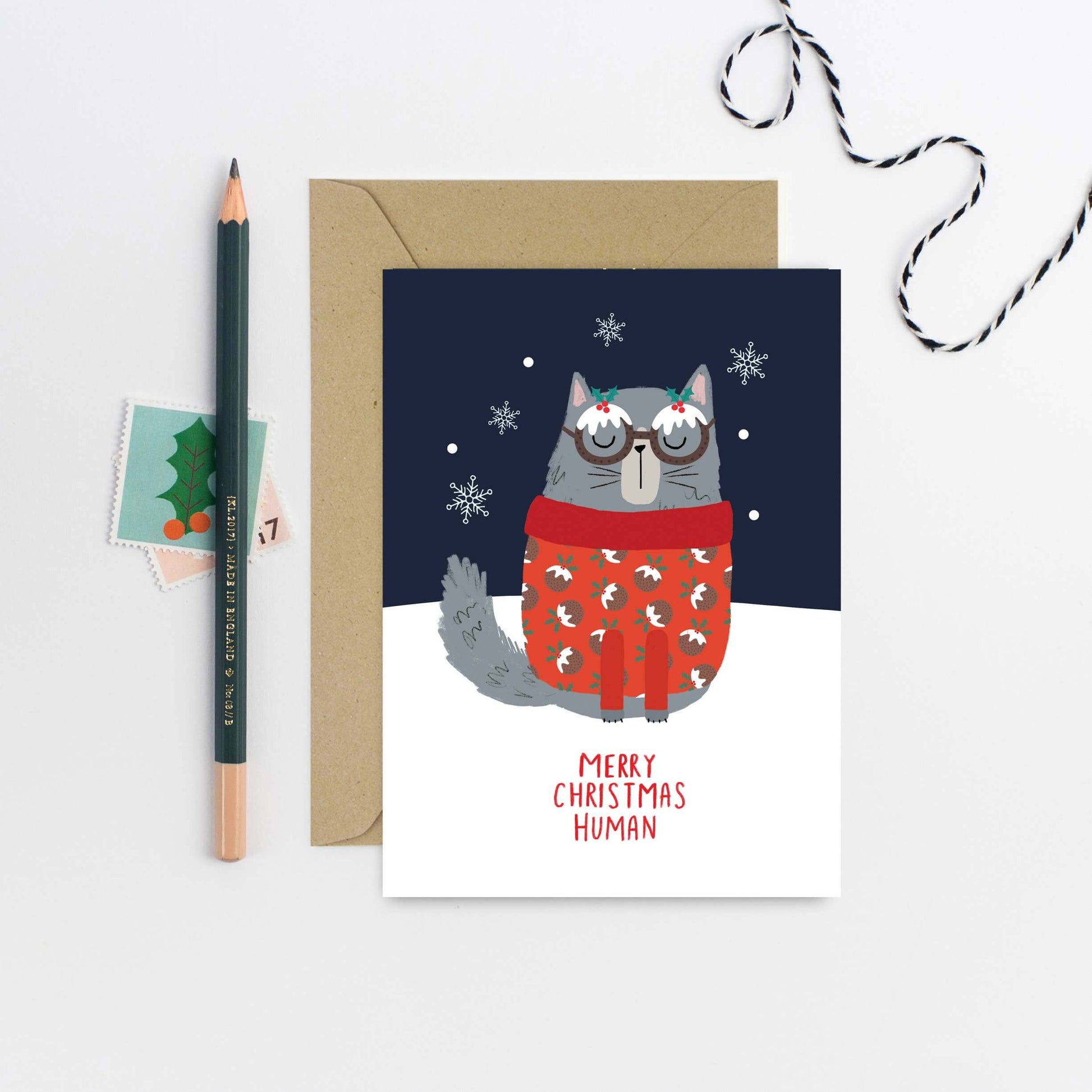 Christmas card with a cat in a sweater and glasses, surrounded by a pencil, envelope, and decorative string.