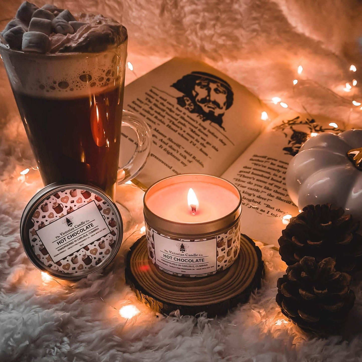 Hot chocolate candle, glass mug with hot chocolate, and book on a soft surface with string lights.
