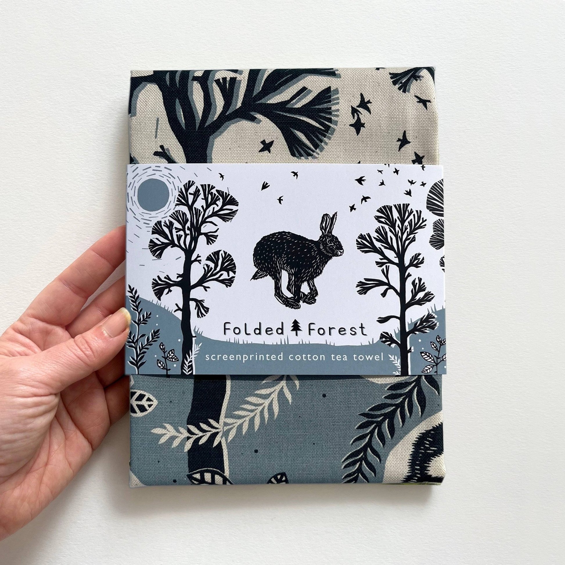 Folded forest-themed cotton tea towel held by a hand on a white background