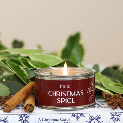 A lit tin candle labeled 'Pintail Christmas Spice' sits on a decorative surface with cinnamon sticks, star anise, and green leaves around it.