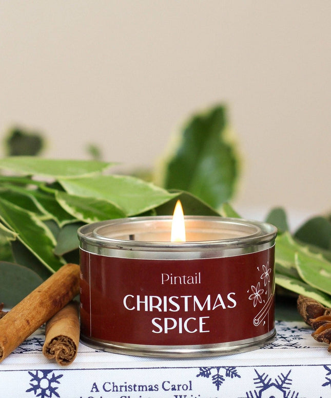 A lit tin candle labeled 'Pintail Christmas Spice' sits on a decorative surface with cinnamon sticks, star anise, and green leaves around it.
