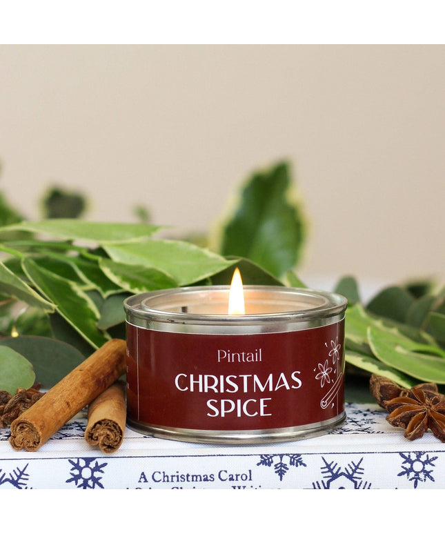 A lit tin candle labeled 'Pintail Christmas Spice' sits on a decorative surface with cinnamon sticks, star anise, and green leaves around it.