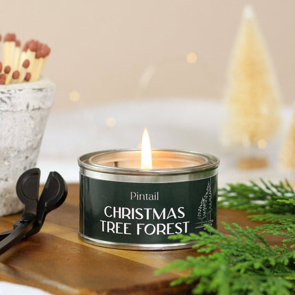 A lit tin candle labeled 'Pintail Christmas Tree Forest' sits on a wooden surface with greenery and a match cup in the background, creating a cozy holiday scene.