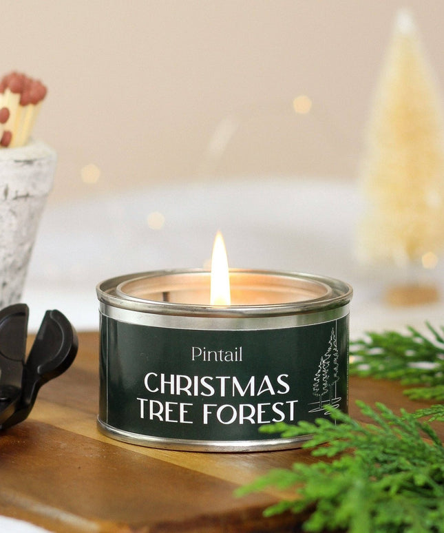 A lit tin candle labeled 'Pintail Christmas Tree Forest' sits on a wooden surface with greenery and a match cup in the background, creating a cozy holiday scene.