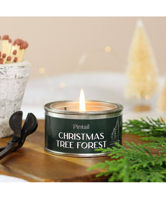 A lit tin candle labeled 'Pintail Christmas Tree Forest' sits on a wooden surface with greenery and a match cup in the background, creating a cozy holiday scene.