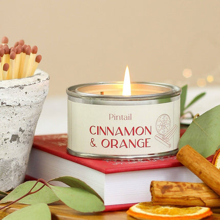 A lit candle in a silver tin labeled 'Pintail Cinnamon & Orange' sits on a red book, surrounded by eucalyptus leaves, cinnamon sticks, dried orange slices, and a small pot of matches against a soft beige background.