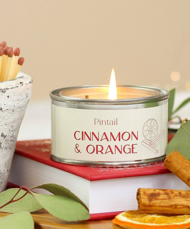 A lit candle in a silver tin labeled 'Pintail Cinnamon & Orange' sits on a red book, surrounded by eucalyptus leaves, cinnamon sticks, dried orange slices, and a small pot of matches against a soft beige background.