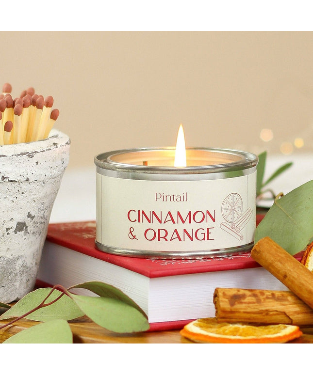 A lit candle in a silver tin labeled 'Pintail Cinnamon & Orange' sits on a red book, surrounded by eucalyptus leaves, cinnamon sticks, dried orange slices, and a small pot of matches against a soft beige background.