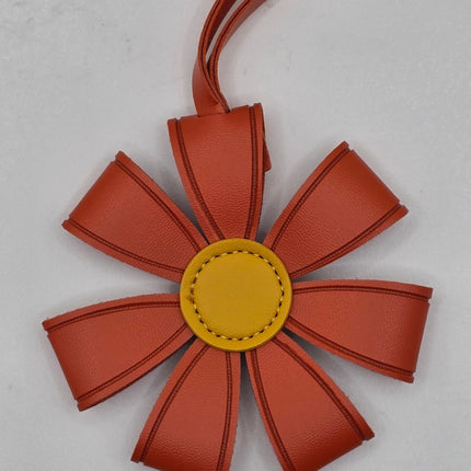 A flower-shaped leather bag charm or luggage tag in terracotta orange with a yellow center, shown on a light gray background.