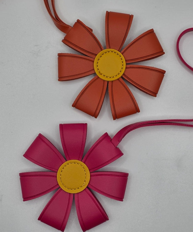 Two flower-shaped leather bag charms or pouches on a light gray background. The top one is terracotta orange with a yellow center, and the bottom one is magenta pink with a yellow center.