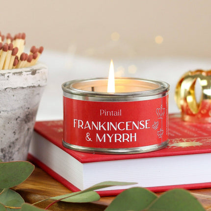 A red scented candle tin labeled 'Pintail FRANKINCENSE & MYRRH' sits on top of a red book, with a lit flame, decorative matches, green leaves, and a softly blurred warm background.