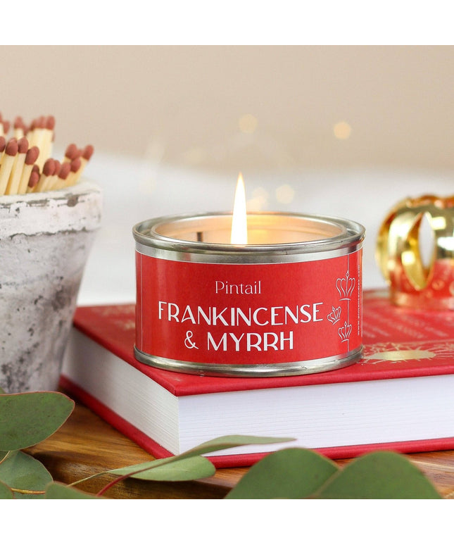 A red scented candle tin labeled 'Pintail FRANKINCENSE & MYRRH' sits on top of a red book, with a lit flame, decorative matches, green leaves, and a softly blurred warm background.