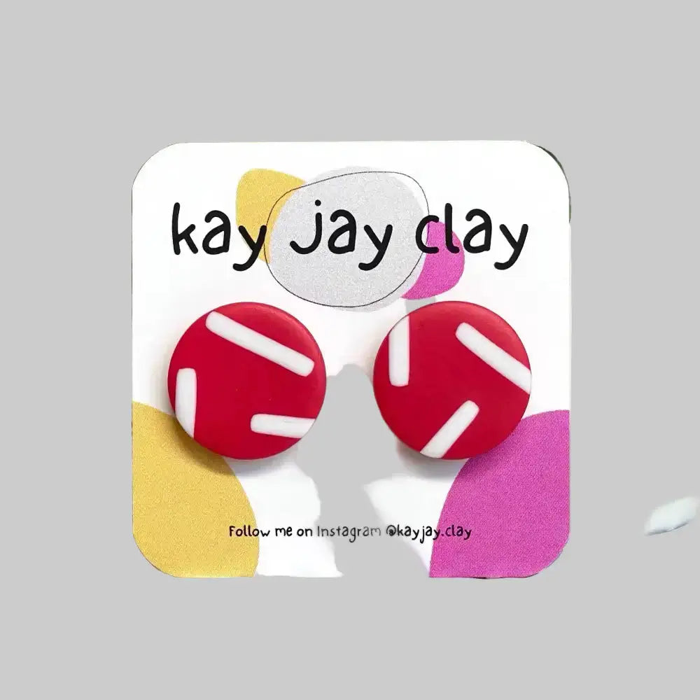 Colorful earrings on a card with 'kay jay clay' branding, set against a plant and colorful surface background.